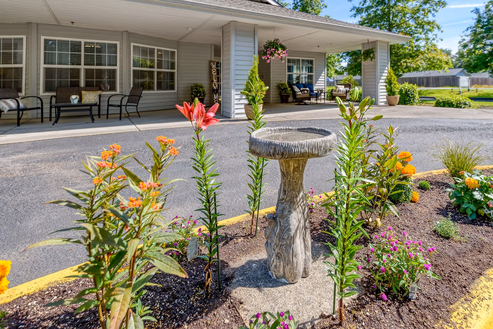 A garden area with colorful flowers and a stone birdbath in the foreground, with a covered porch featuring outdoor seating and a welcome sign in the background at Creekside Place Memory Care.