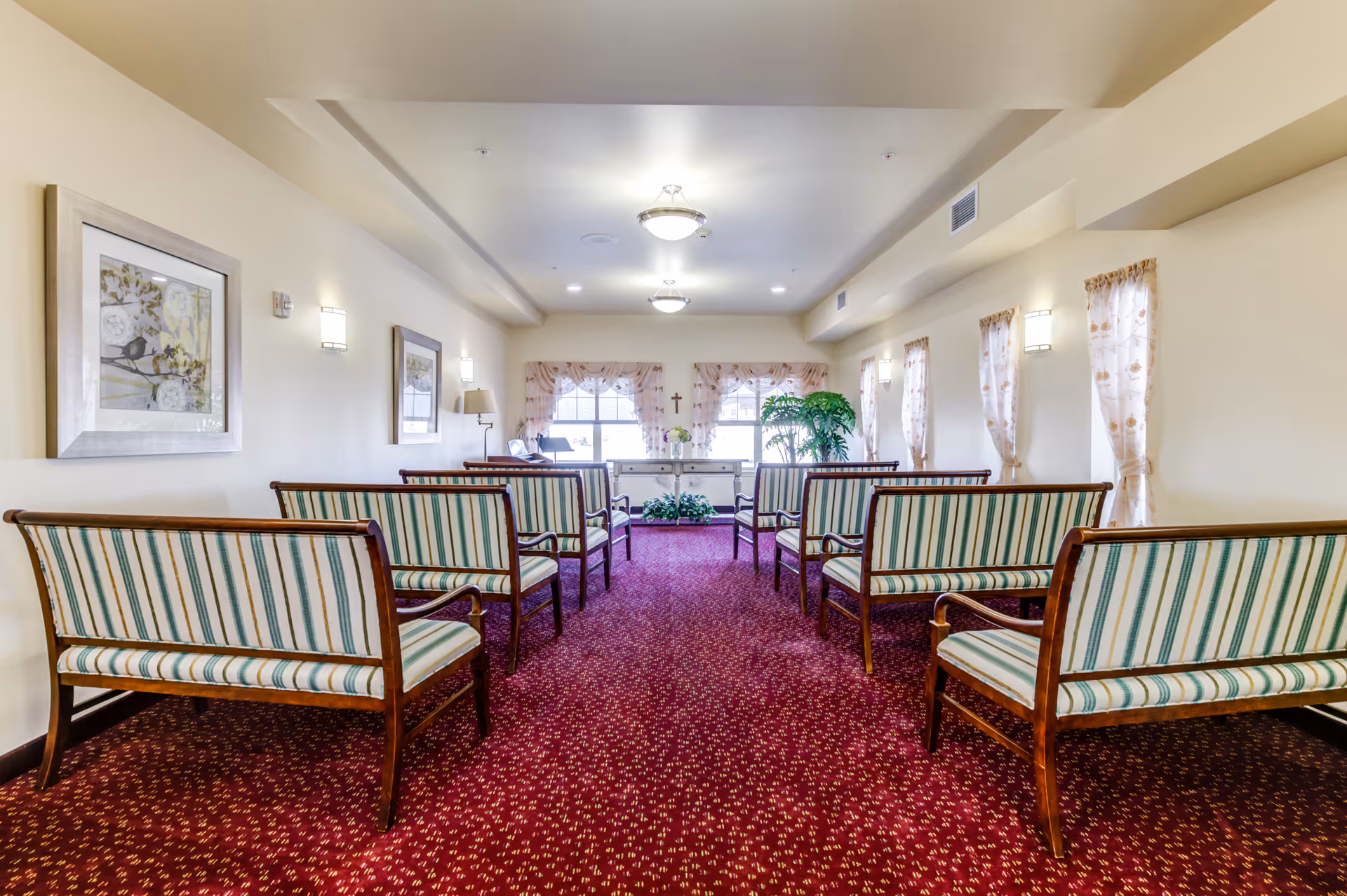 A quiet room with rows of wooden benches upholstered in green and white striped fabric, arranged facing a small table with floral decorations and a cross on the wall behind it. The room has cream-colored walls, framed artwork, patterned red carpet, and windows with sheer curtains letting in natural light.