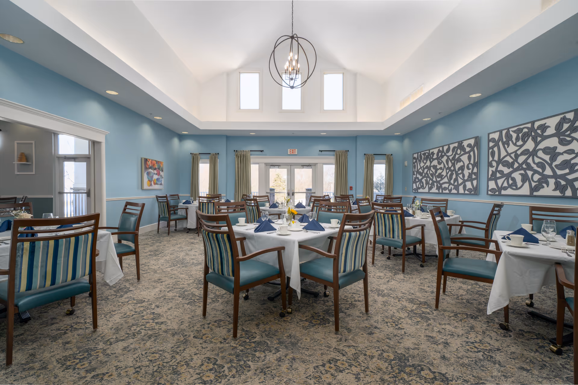 A bright dining room with blue walls and a high ceiling featuring three tall windows. Several tables are set with white tablecloths, blue napkins, cups, and glasses. Wooden chairs with blue and green striped cushions surround the tables. Decorative wall art and a modern chandelier add to the elegant atmosphere.