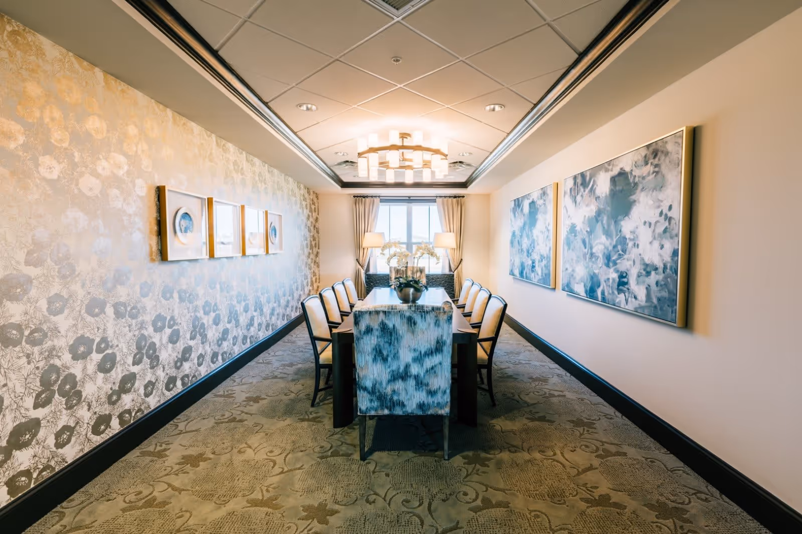 A long dining room with a rectangular table surrounded by ten chairs, two large abstract paintings on the right wall, three framed artworks on the left wall with floral wallpaper, a modern chandelier hanging from the ceiling, and a window with curtains at the far end letting in natural light.