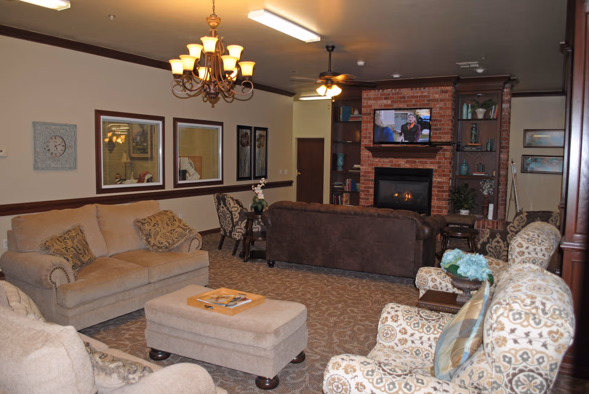 A cozy senior living community lounge area with beige and patterned armchairs, a beige sofa, a brown sofa, and a matching ottoman. The room features a brick fireplace with a TV mounted above it, built-in wooden shelves with decorative items, a chandelier, and framed artwork on the walls.