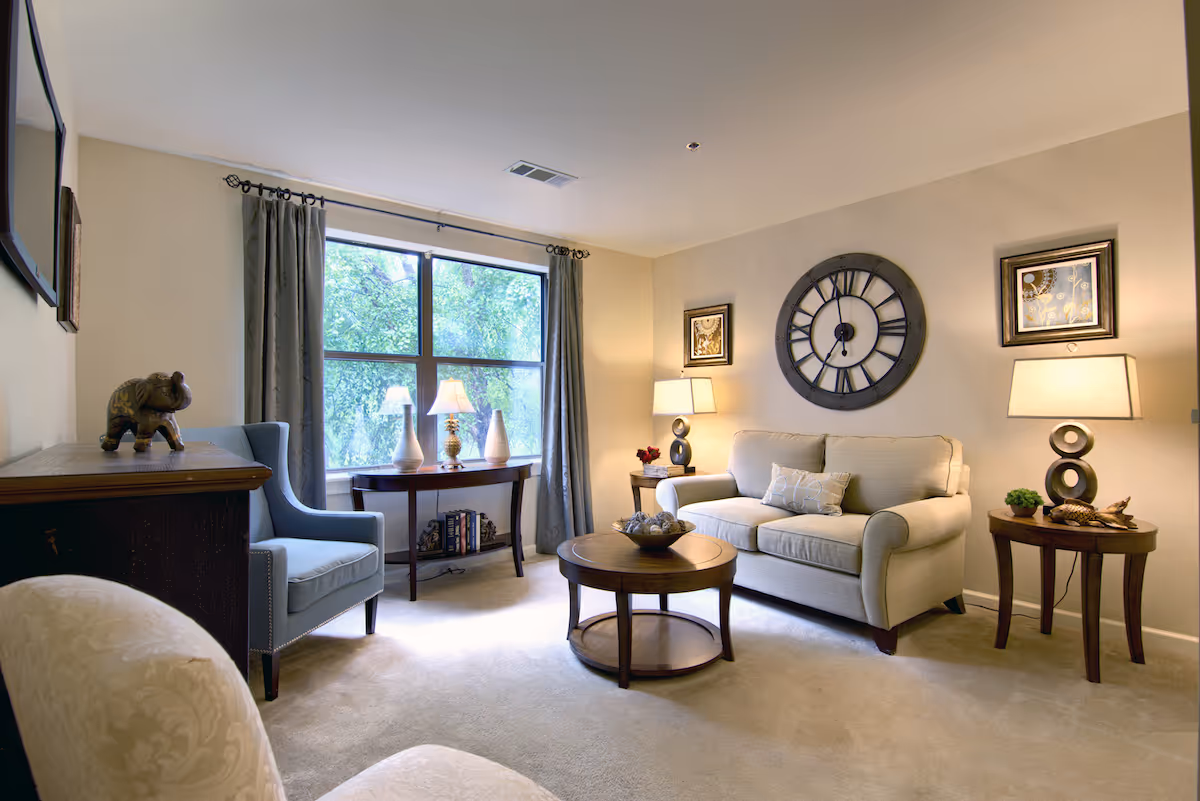 A cozy living room with a beige sofa, a round wooden coffee table, two side tables with lamps, a large wall clock, framed artwork, a blue armchair, a wooden console table with a decorative elephant, and a large window with curtains letting in natural light.