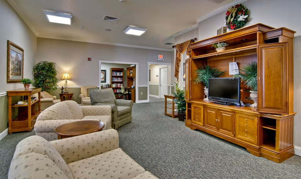 A cozy senior living facility common area with beige patterned armchairs and sofas arranged around a wooden coffee table. A large wooden entertainment center with a flat-screen TV and decorative plants is on the right side. The room has carpeted floors, soft lighting from table lamps, framed artwork on the walls, and an open doorway leading to a room with bookshelves.