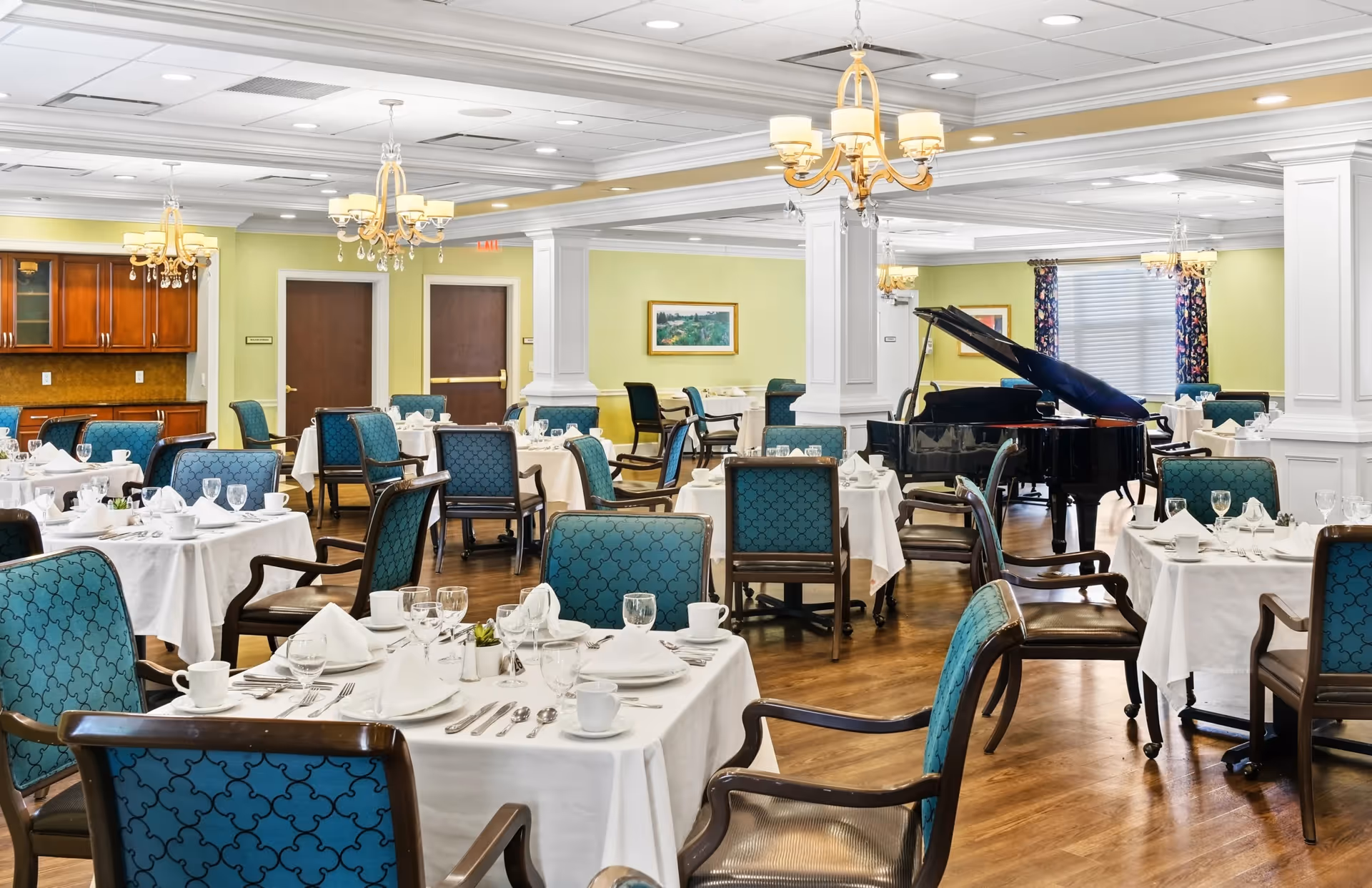 A spacious dining room with multiple tables covered in white tablecloths, set with plates, cups, glasses, and silverware. The chairs have blue patterned upholstery and wooden frames. The room features chandeliers hanging from the ceiling, a grand piano in the background, light green walls, and wooden flooring.