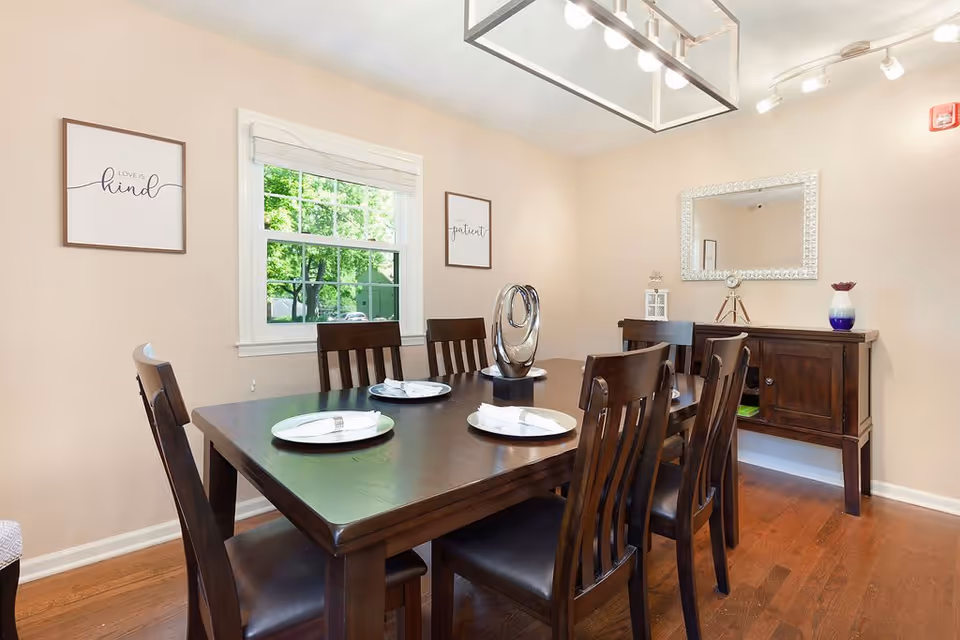 A dining room with a dark wooden table set for four with white plates and silverware. Six matching wooden chairs surround the table. On the table is a modern silver decorative sculpture. The room has light beige walls, a window with white trim showing green trees outside, and two framed wall art pieces with the words 'Love is kind' and 'Be patient'. A wooden sideboard with decorative items and a large mirror above it is against the far wall. The floor is hardwood, and the ceiling has track lighting and a rectangular pendant light fixture.