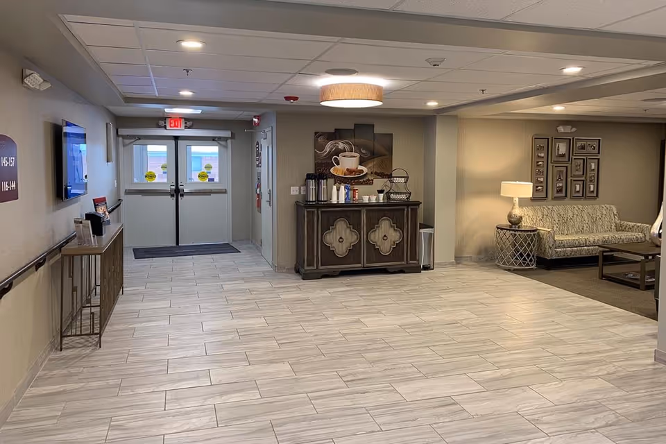 Interior view of a senior living facility hallway with a coffee station against the wall, a seating area with a patterned sofa, side table with lamp, and framed pictures on the wall. Double exit doors with windows are visible at the end of the hallway.