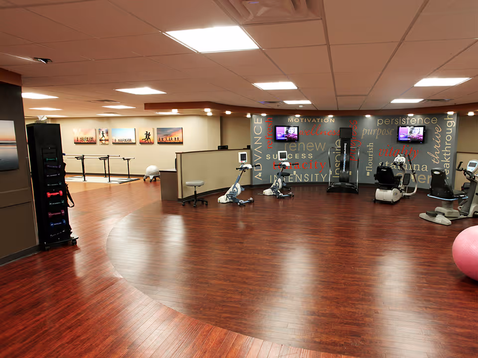 A spacious fitness room with wooden flooring and exercise equipment including stationary bikes and a treadmill. The walls are decorated with motivational words and several framed pictures of people running. There is also a rack with colorful dumbbells and a large pink exercise ball on the floor.