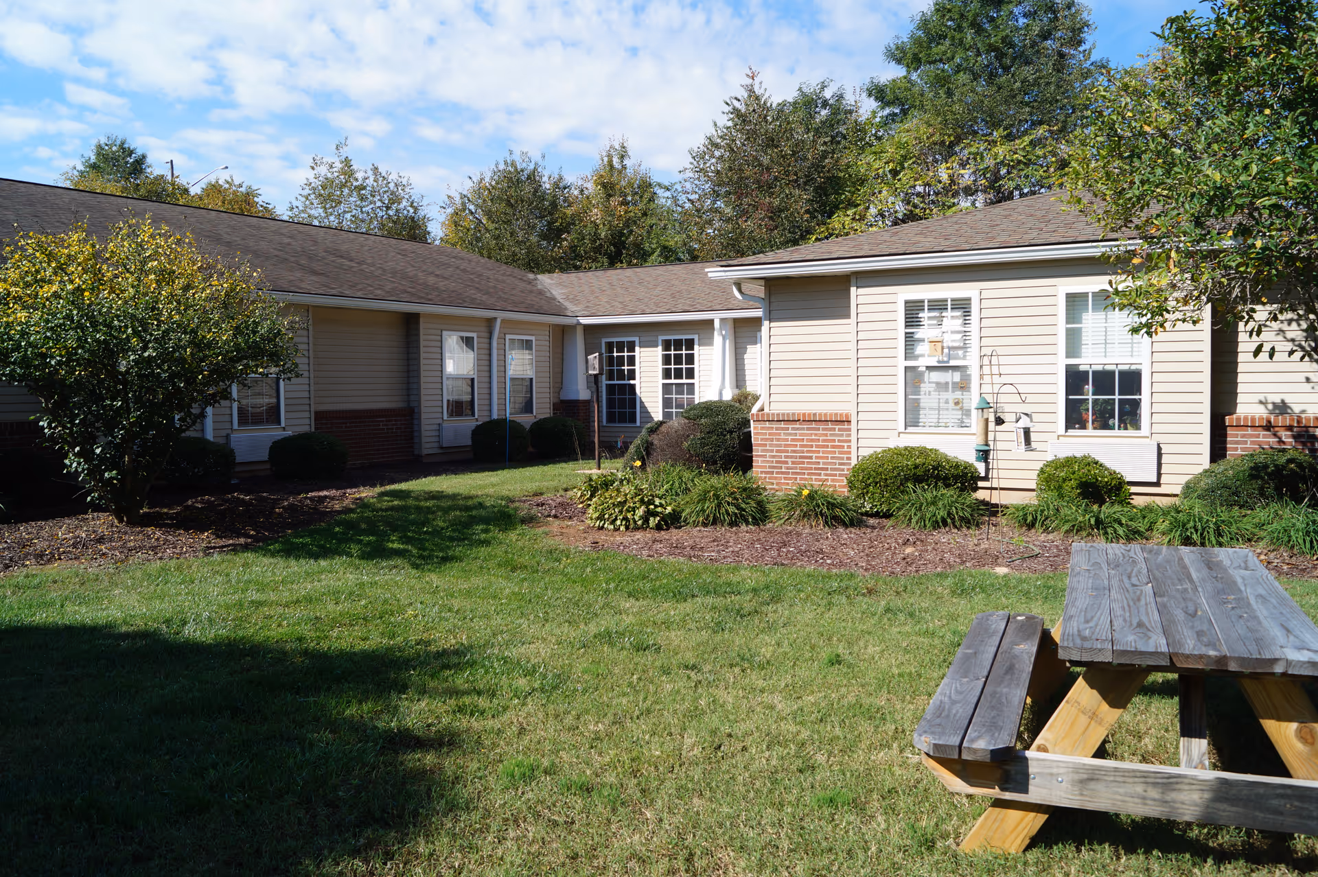 Beige single-story building forming a courtyard with shrubs and a wooden picnic table on a grassy lawn.