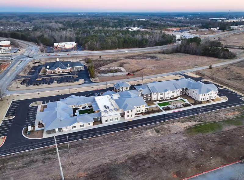 Aerial view of a large senior living facility named Canopy at Westridge, showing a modern, multi-wing building with a gray roof surrounded by parking lots and roads, with some trees and open land in the background.