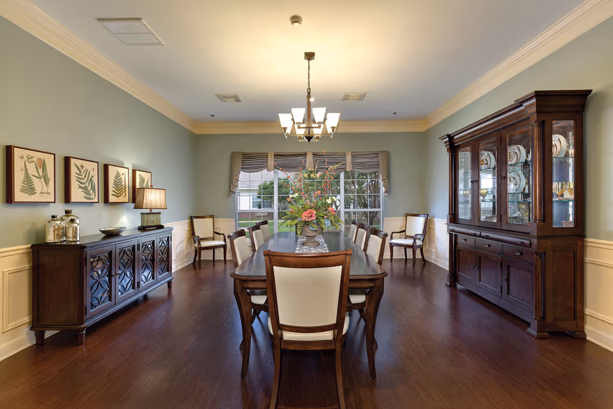 A formal dining room with a long wooden table surrounded by eight upholstered chairs. A large floral centerpiece is on the table. The room features wooden flooring, light green walls with white wainscoting, a chandelier hanging from the ceiling, a wooden sideboard with decorative items and framed botanical prints on the left wall, and a large wooden china cabinet displaying plates and glassware on the right. A window with valances provides natural light at the far end of the room.