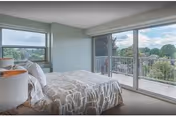 A bright bedroom with a large bed covered in a patterned beige and white bedspread, two bedside tables with lamps, a large window on the left, and sliding glass doors leading to a balcony with a view of trees and a partly cloudy sky.
