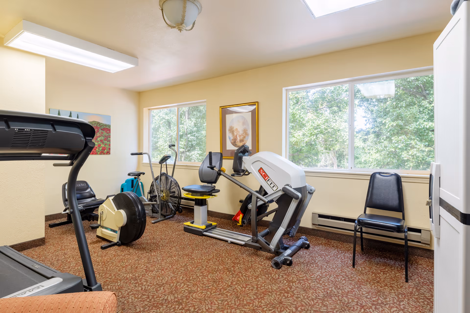 A small fitness room in a senior living community with exercise equipment including a treadmill, rowing machine, stationary bike, and a recumbent bike. The room has beige walls, two large windows showing green trees outside, a patterned carpet, and a black chair against the wall. There are two framed pictures on the walls.