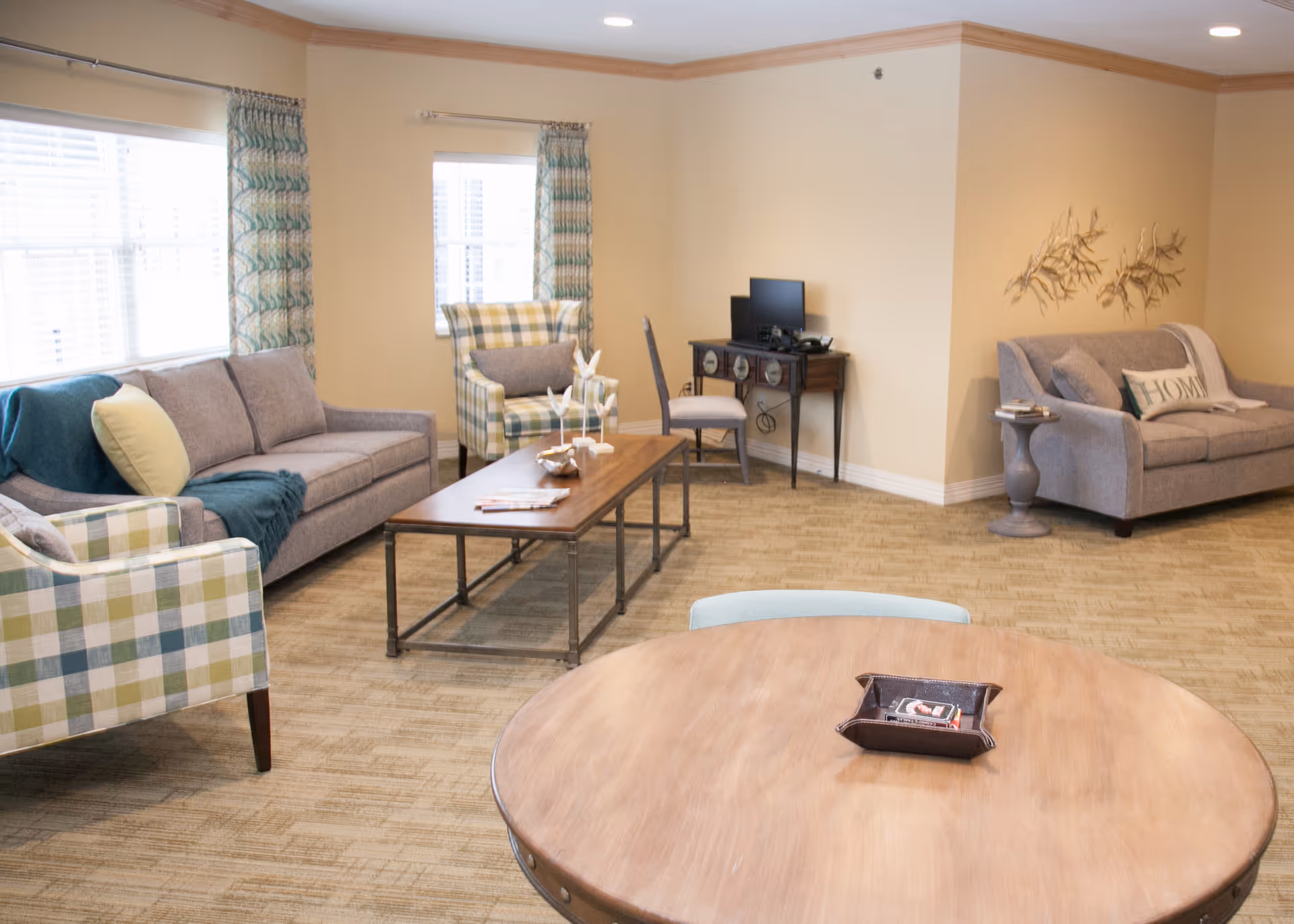 A cozy living room with beige walls and carpeted floor. The room features two gray sofas, two plaid armchairs, a wooden coffee table with decorative items, a round wooden table with a tray, and a small desk with a computer and chair. The windows have patterned curtains, and there are decorative wall hangings above one sofa.