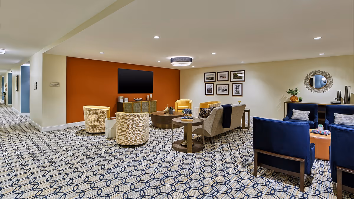 A spacious senior living facility common area with patterned carpet flooring, beige walls, and recessed lighting. The room features a seating arrangement including yellow and blue armchairs, a beige sofa, round wooden coffee tables, a flat-screen TV mounted on an orange accent wall, framed pictures, and decorative items such as plants and a mirror.