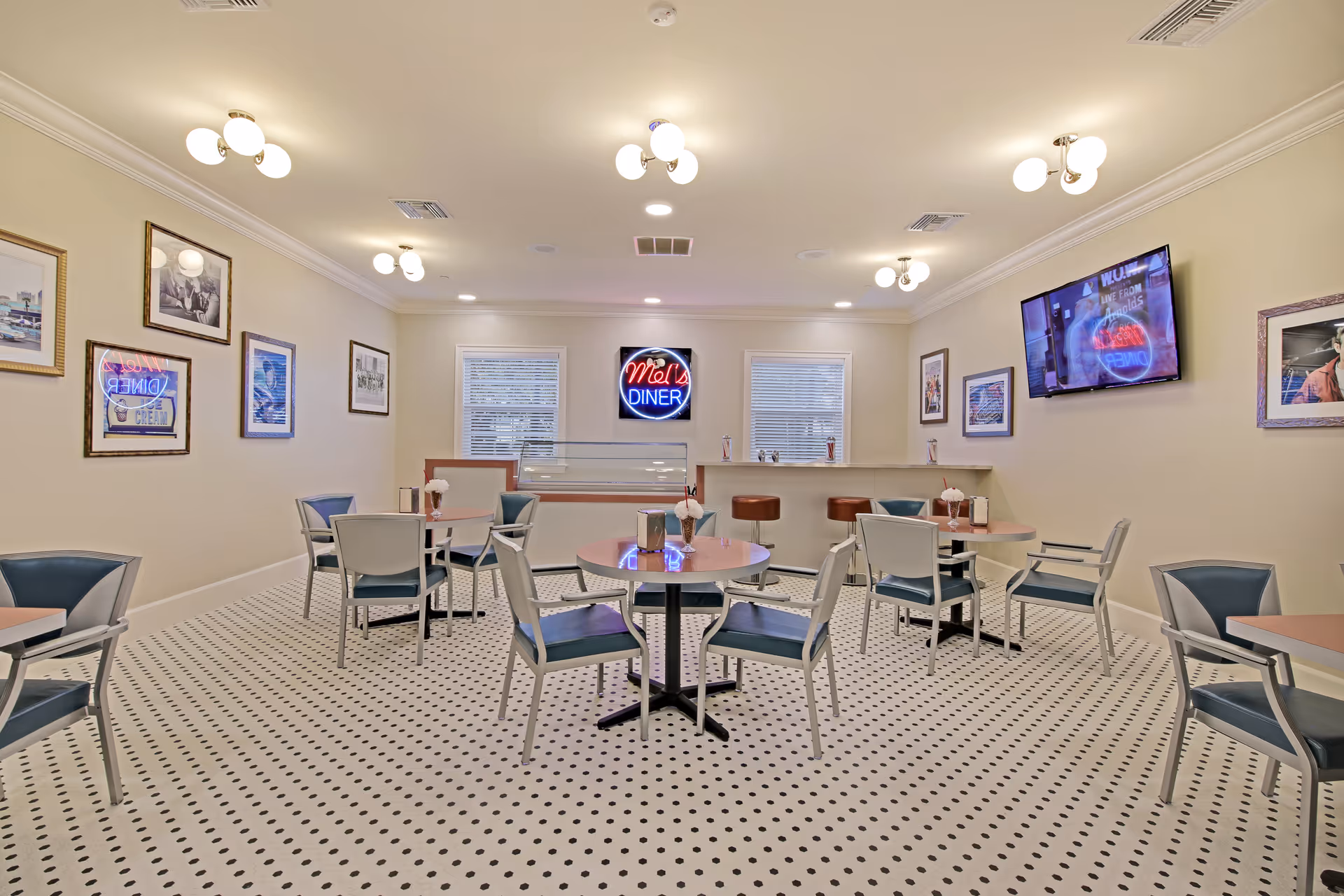 A bright dining room with round tables and chairs arranged neatly on a white floor with black dots. The walls are decorated with framed pictures and a neon sign that reads 'Mel's Diner'. There is a counter with bar stools and two windows with blinds. A flat-screen TV is mounted on the right wall.