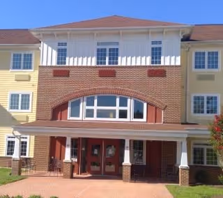 Front exterior view of a multi-story senior living facility building with a combination of brick and light yellow siding, large windows, and a covered entrance with columns.