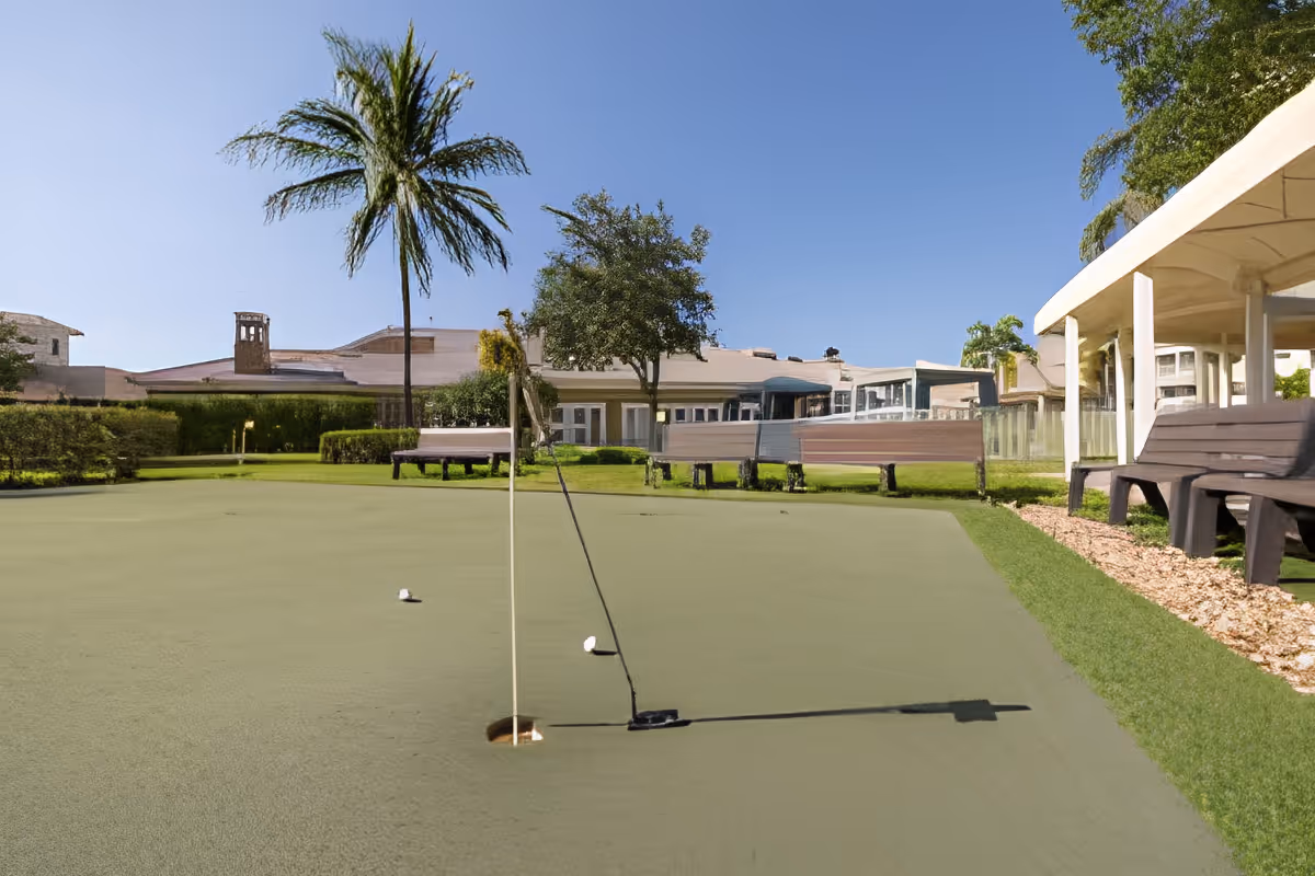 Outdoor putting green with a flagstick, golf balls, benches, a palm tree and clubhouse buildings under a clear sky.