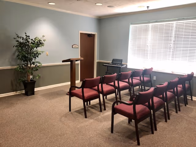 A small meeting or presentation room with two rows of red cushioned chairs facing a wooden podium. There is a potted plant in the corner, a keyboard on a stand near a large window with blinds, and a closed door on the far wall. The walls are painted light blue and green with white trim.