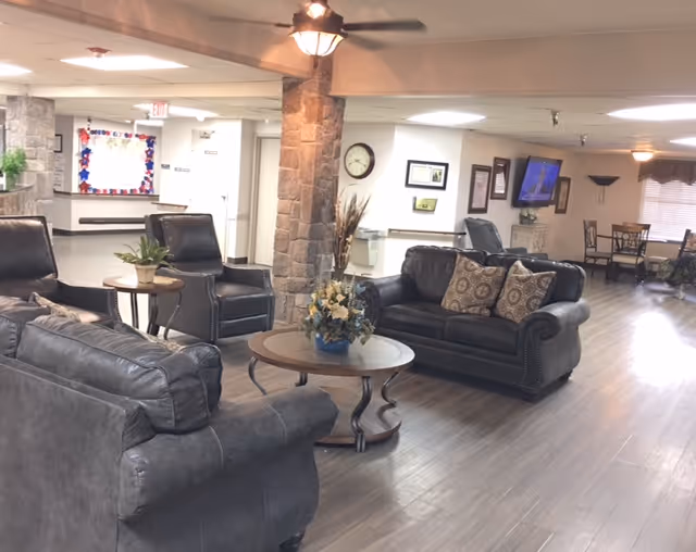 A spacious and well-lit common area in a nursing and rehabilitation center featuring dark leather sofas and armchairs arranged around a wooden coffee table with a floral centerpiece. The room has wood flooring, a ceiling fan, a stone column, a wall clock, a mounted TV, and a dining area with tables and chairs in the background.