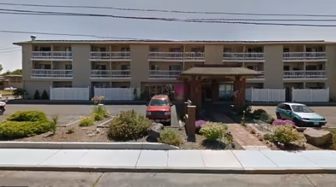 Front exterior of a three-story senior care building with balconies, a covered entrance canopy, parked cars, and landscaped beds.
