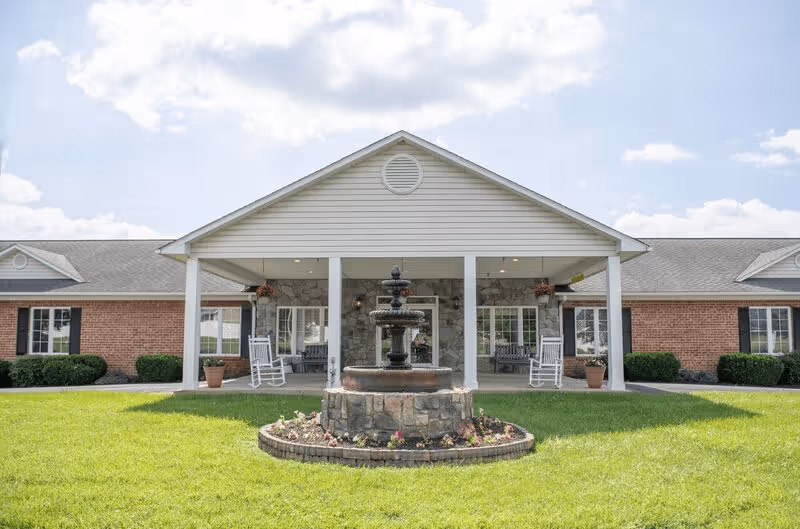 Front exterior view of a single-story senior living facility building with a covered porch featuring white rocking chairs and a central stone water fountain surrounded by a flower bed on a green lawn under a partly cloudy sky.