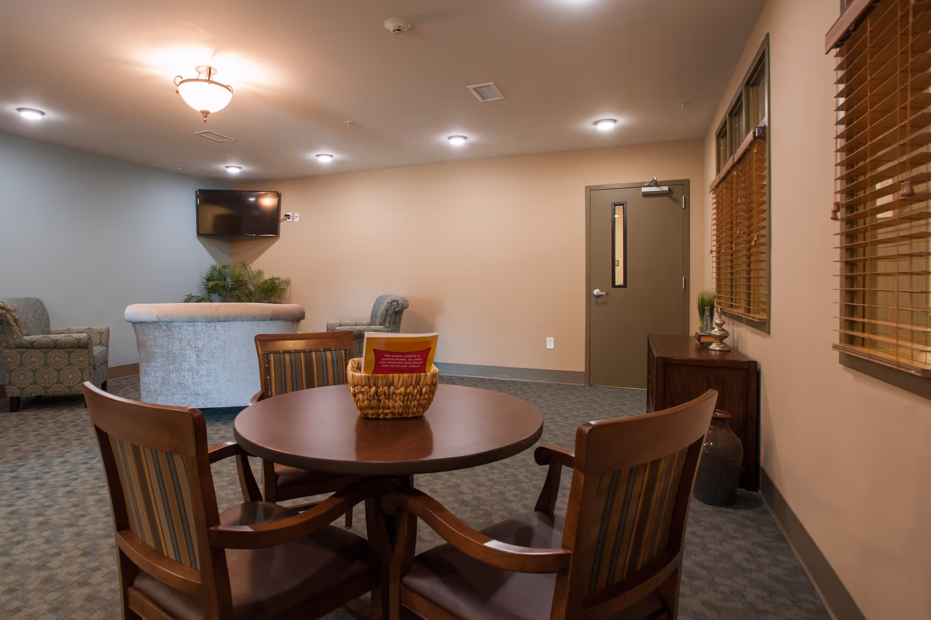 A cozy senior living common area with a round wooden table and four chairs in the foreground. In the background, there are two upholstered armchairs and a curved sofa facing a wall-mounted flat screen TV. The room has beige walls, carpeted floor, a closed door, and windows with wooden blinds on the right side. A small basket with a red card is on the table, and a wooden cabinet with decorative items is against the wall.