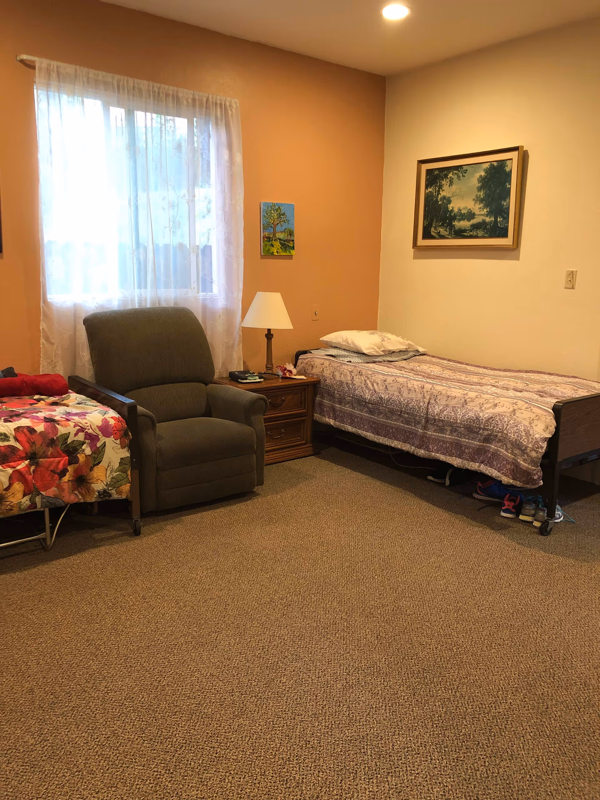 A cozy bedroom with two single beds, one with a floral bedspread and the other with a patterned purple and white bedspread. Between the beds is a wooden nightstand with a lamp and some small items. There is a comfortable armchair next to the window, which has sheer white curtains. The walls are painted in two tones, peach and cream, and two framed paintings hang on the walls. The floor is carpeted in a neutral color.