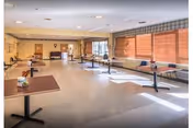 A spacious, well-lit room with multiple tables and chairs arranged along the walls and center, large windows with blinds allowing natural light, and a clean, open floor space suitable for group activities or dining.