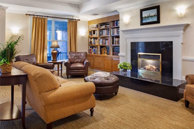 A cozy living room with a beige sofa, two brown leather armchairs, a round leather ottoman, a fireplace with a black marble hearth, a built-in wooden bookshelf filled with books, a side table with a lamp, and yellow curtains on the window.