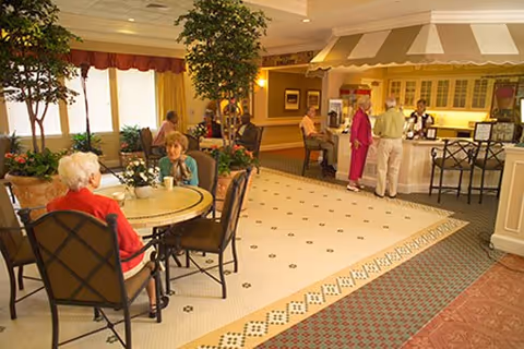 Residents seated at round tables in a bright dining/cafe area with a service counter and indoor plants.