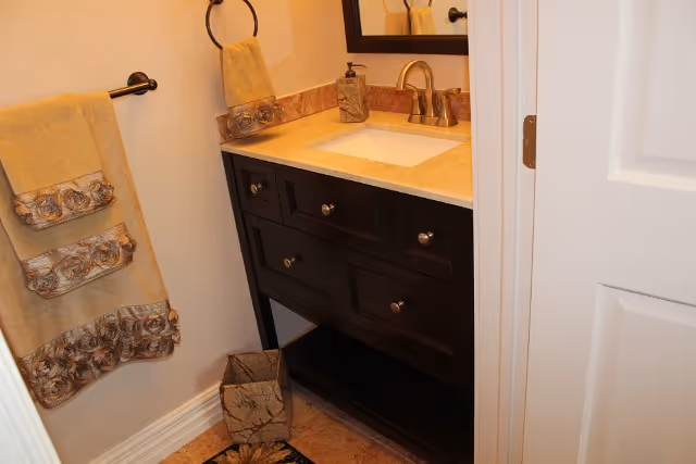 A small bathroom vanity area with a dark wood cabinet, beige countertop, and a built-in sink with a silver faucet. A mirror is mounted on the wall above the sink. On the left wall, there is a towel rack holding two beige towels with decorative floral trim. A matching hand towel hangs on a ring above the countertop. A small wastebasket is placed on the floor next to the vanity.