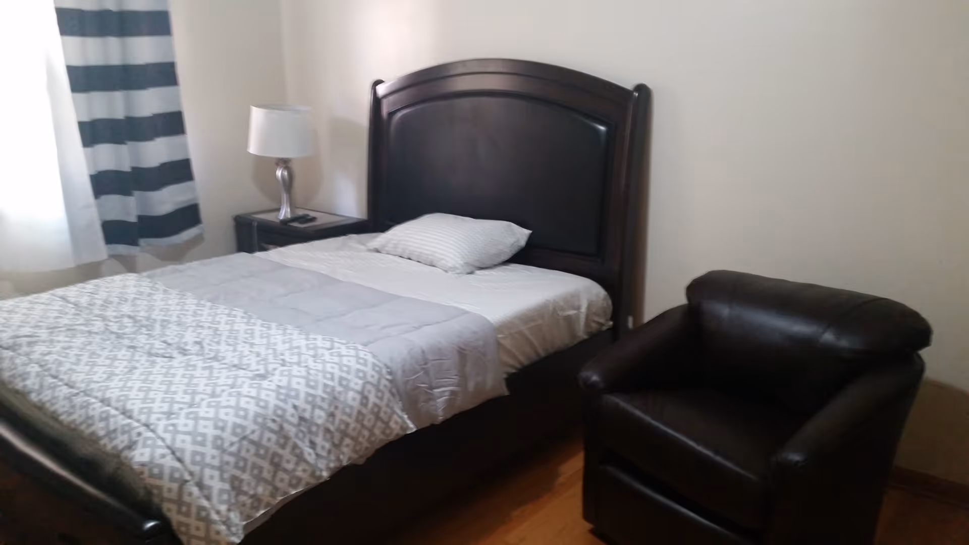 A bedroom with a dark wooden bed frame, a bed made with a white and gray patterned comforter and a single pillow. Next to the bed is a nightstand with a silver lamp and a remote control. A dark brown leather armchair is positioned near the bed. The room has light-colored walls and a window with white and dark gray striped curtains.