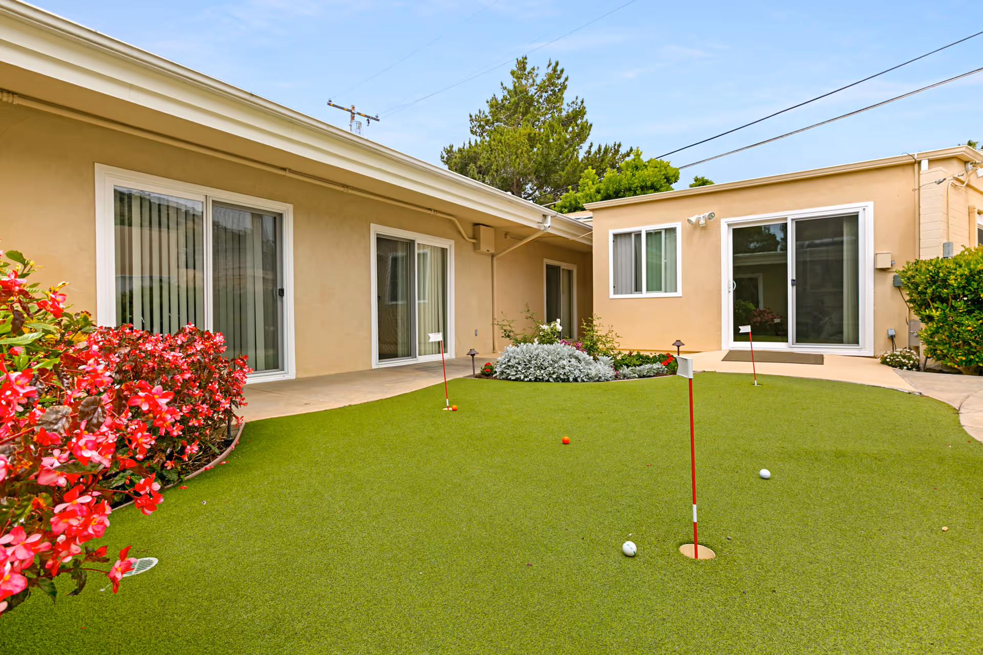 Courtyard with a small putting green, red flowers, and sliding glass doors of a beige senior living building.