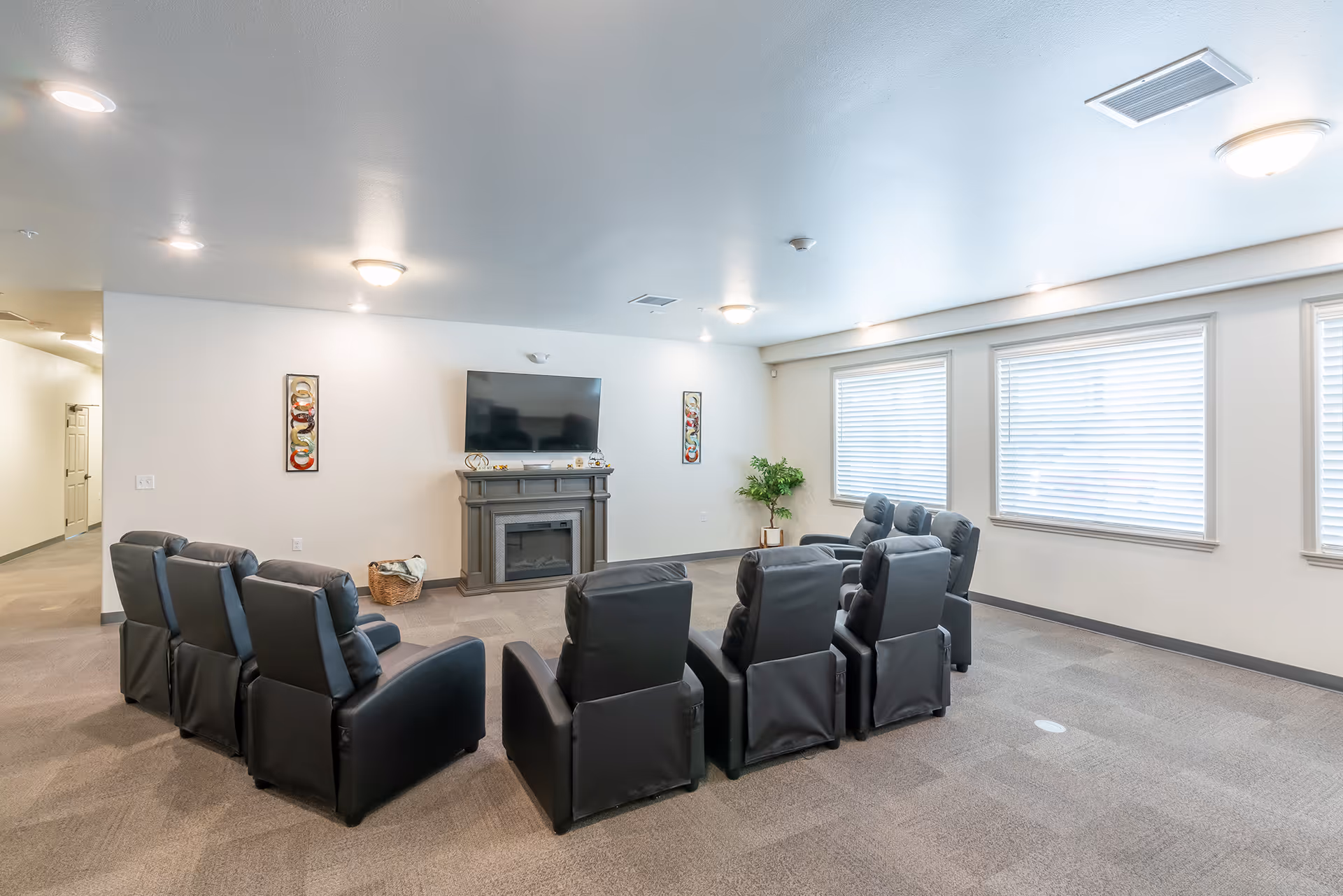 A spacious living room with a semi-circle arrangement of black recliner chairs facing a wall-mounted flat-screen TV above a gray electric fireplace. The room has beige walls, carpeted floor, three large windows with blinds, ceiling lights, and a small green plant in the corner.