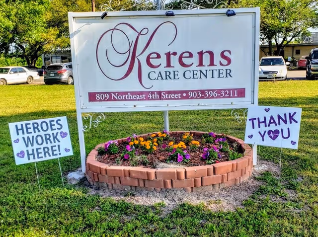Outdoor sign for Kerens Care Center with address 809 Northeast 4th Street and phone number 903-396-3211, surrounded by a circular flower bed with colorful flowers. Two smaller signs on either side read 'HEROES WORK HERE!' and 'THANK YOU' with purple hearts.