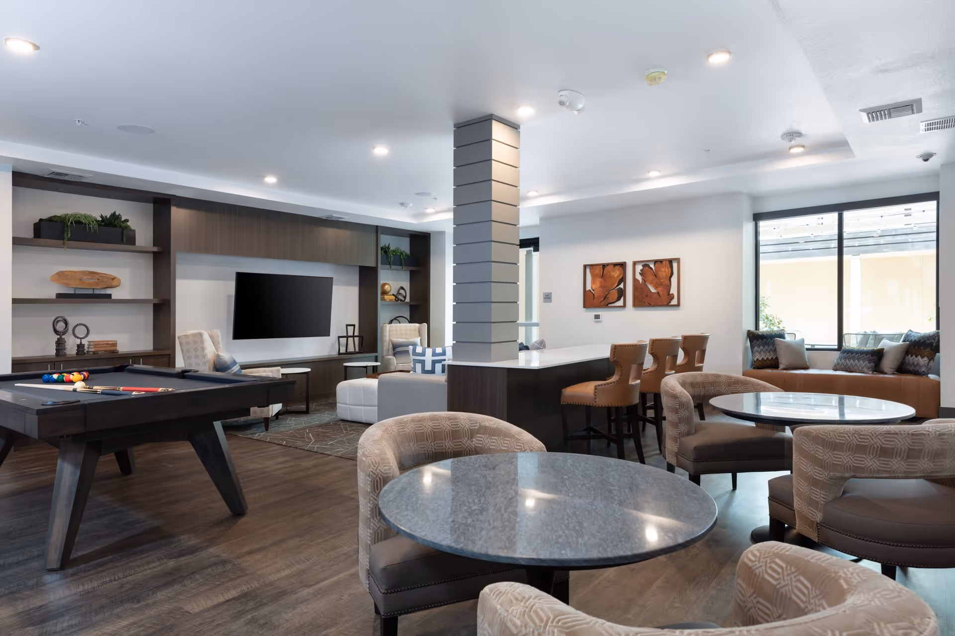 A modern and spacious common area in Santana Terrace featuring a pool table, round tables with upholstered chairs, a large TV mounted on a dark wood entertainment unit, and a seating area with a long cushioned bench by the window. The room has wood flooring, recessed lighting, and decorative wall art.