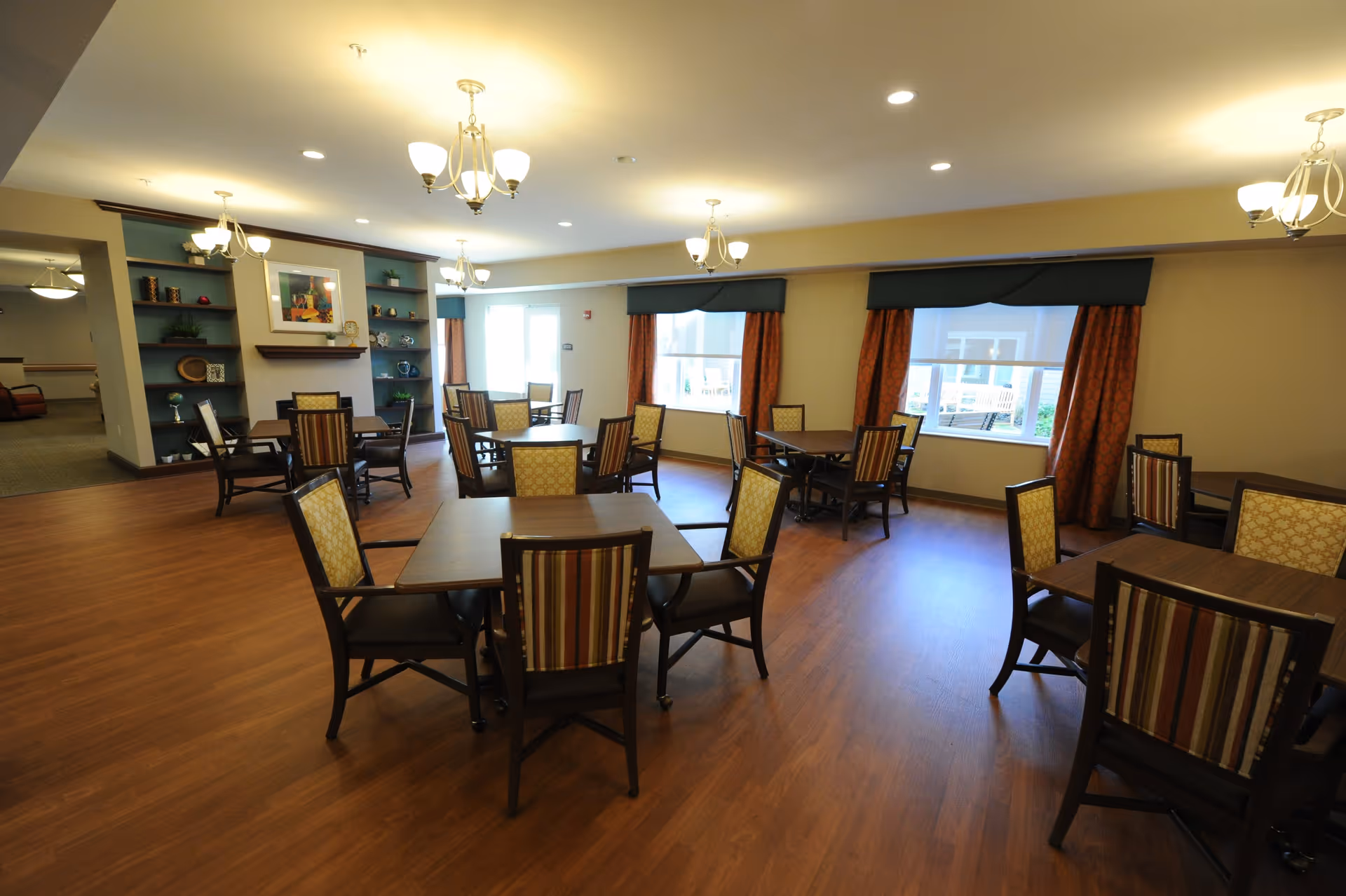 A spacious dining room with multiple square wooden tables and cushioned chairs arranged neatly. The room has large windows with orange curtains and valances, wooden flooring, and several ceiling light fixtures. There is a built-in shelving unit with decorative items and a framed picture on the far wall.