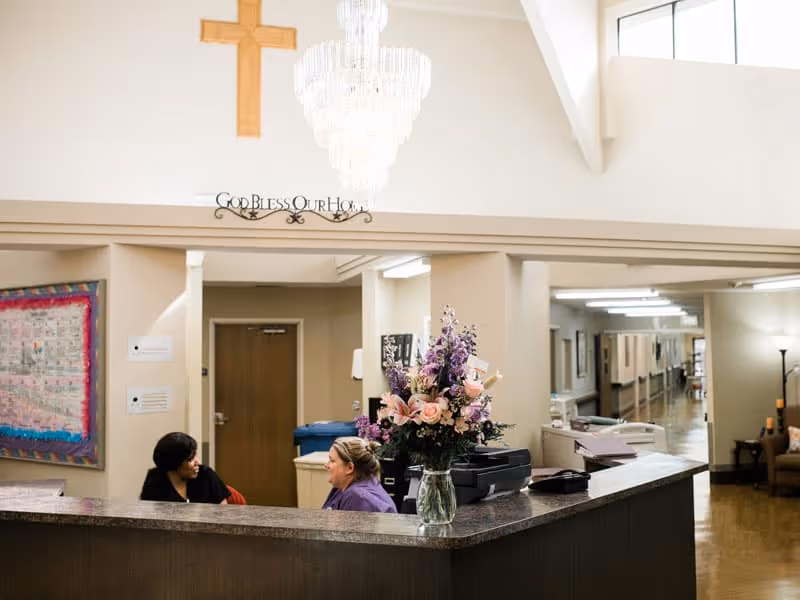 Reception area inside a nursing and rehab facility with two staff members talking behind the front desk. A large floral arrangement is on the desk. Above the desk is a chandelier and a wooden cross on the wall, with a decorative sign that reads 'God Bless Our House'. A hallway extends to the right with seating and lamps visible.