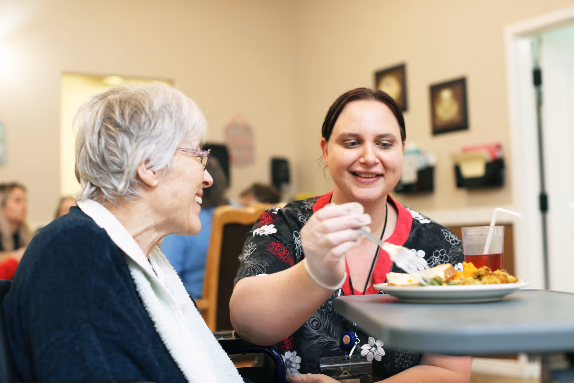 A caregiver feeds a smiling elderly woman at a dining table in a memory care facility.