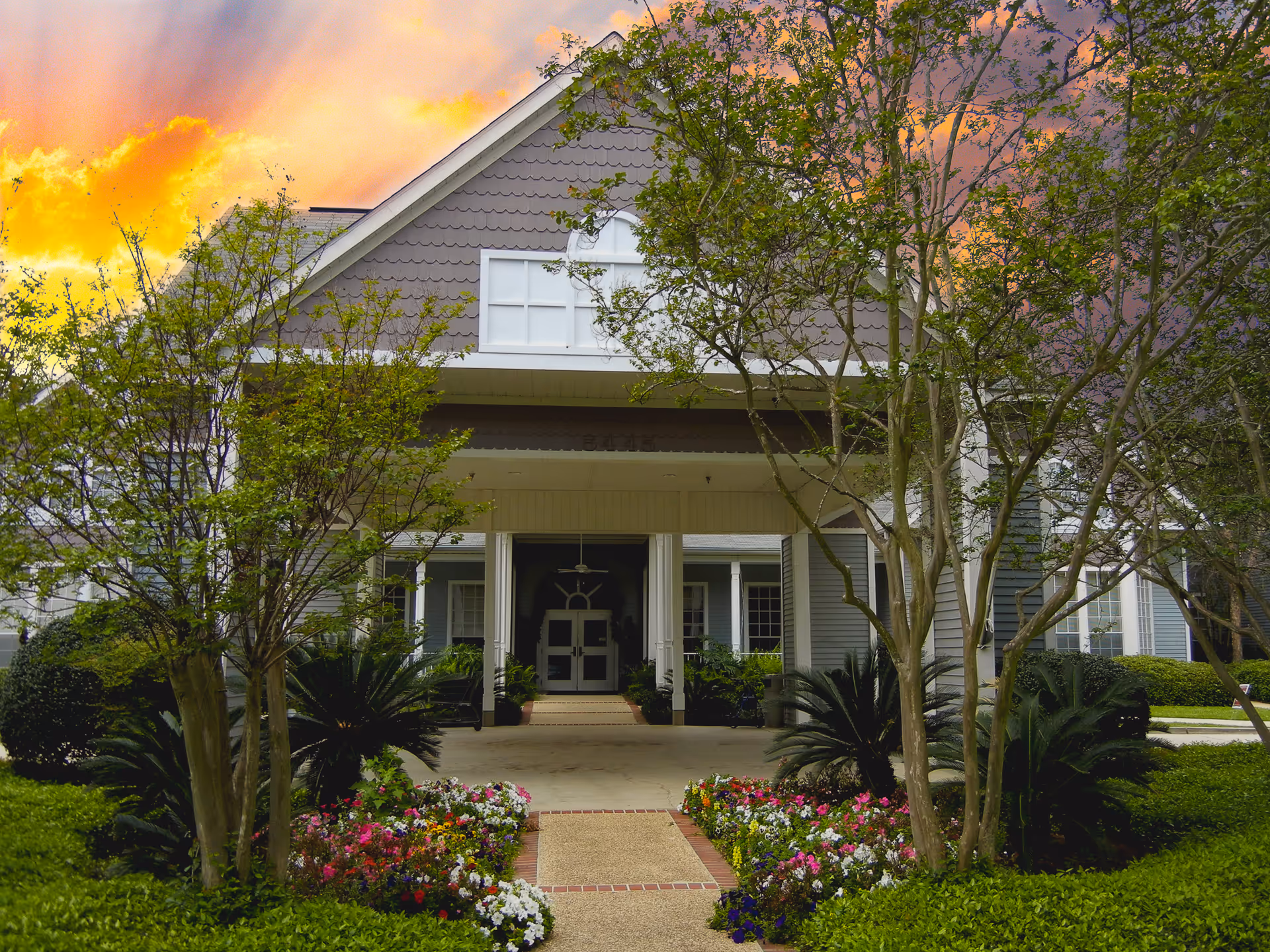 Front exterior view of Williamsburg Senior Living Community building with a covered entrance, surrounded by trees and colorful flower beds under a vibrant sunset sky.