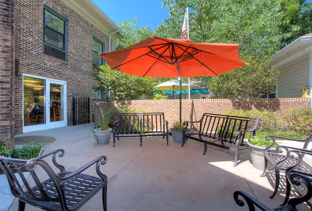 Outdoor patio area with black metal benches and chairs arranged around a large red umbrella. The patio is adjacent to a brick building with glass doors and windows. There are potted plants and a brick wall enclosing the space, with trees and greenery visible beyond the wall under a clear blue sky.