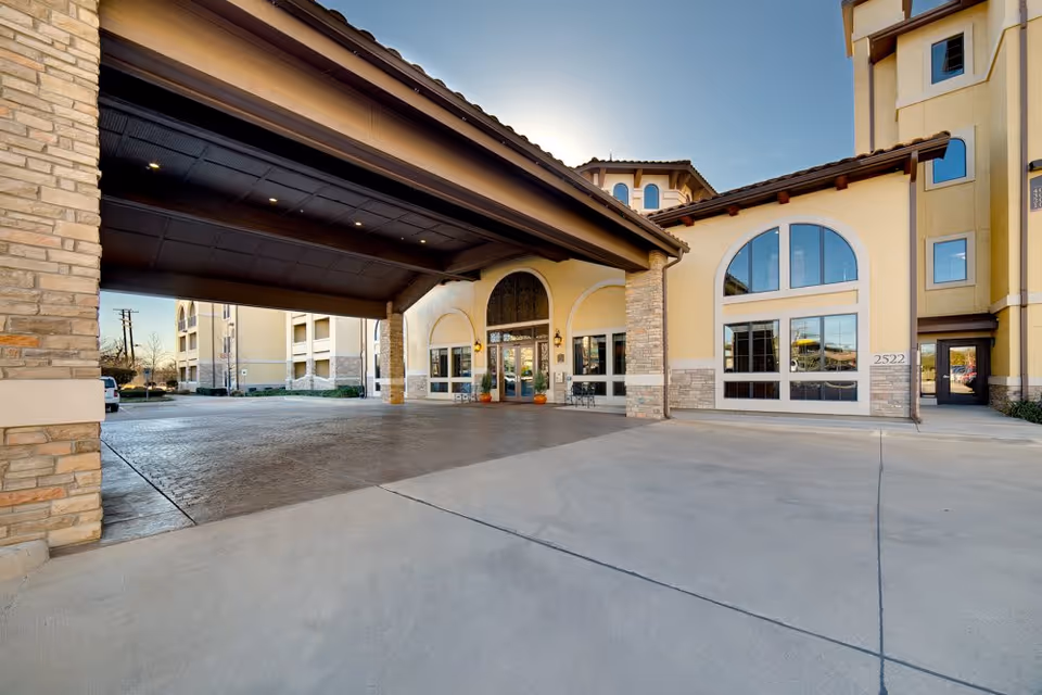 Exterior view of Aventine At Kessler Park showing the entrance with a covered driveway, large arched windows, and a beige and stone facade under a clear sky.