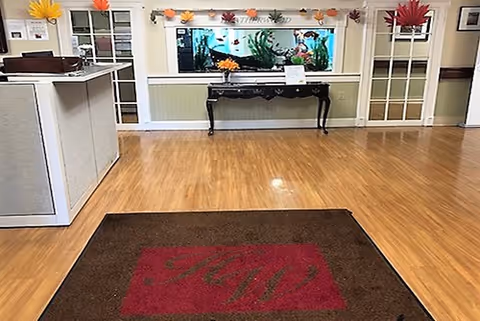 Interior view of a facility lobby or reception area with wooden flooring, a brown mat with red initials, a black table against the wall holding a flower arrangement and a sign, and a large aquarium built into the wall. There are decorative autumn leaves on the walls and glass-paneled doors on either side of the aquarium.