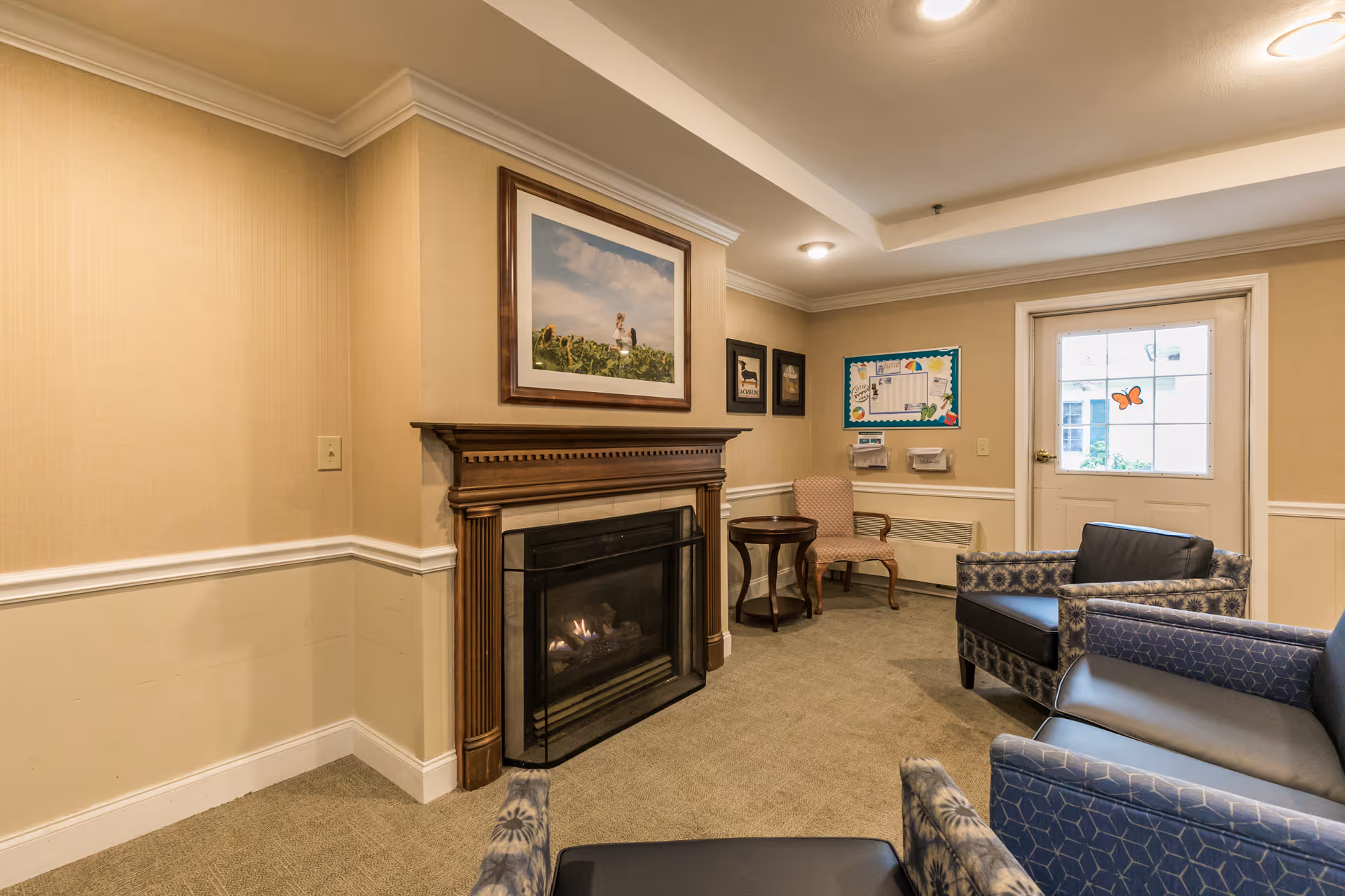 Cozy lounge with a fireplace, upholstered chairs, small tables, wall art, and a door leading outside.