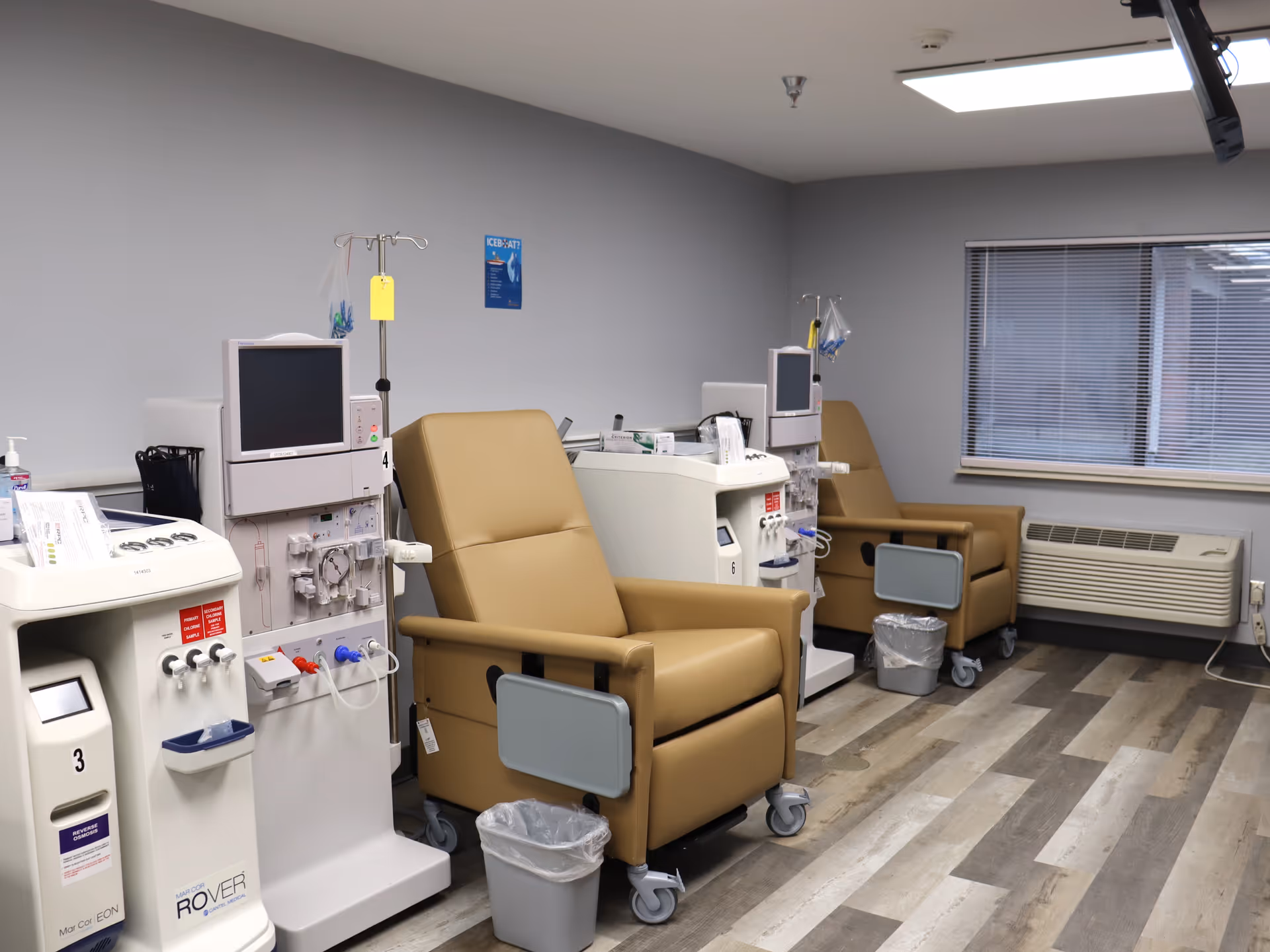 A medical treatment room with two beige recliner chairs equipped with dialysis machines. The room has a window with blinds, a wall-mounted TV, and a light-colored wood floor. There are small trash bins next to each chair and medical equipment positioned beside the chairs.