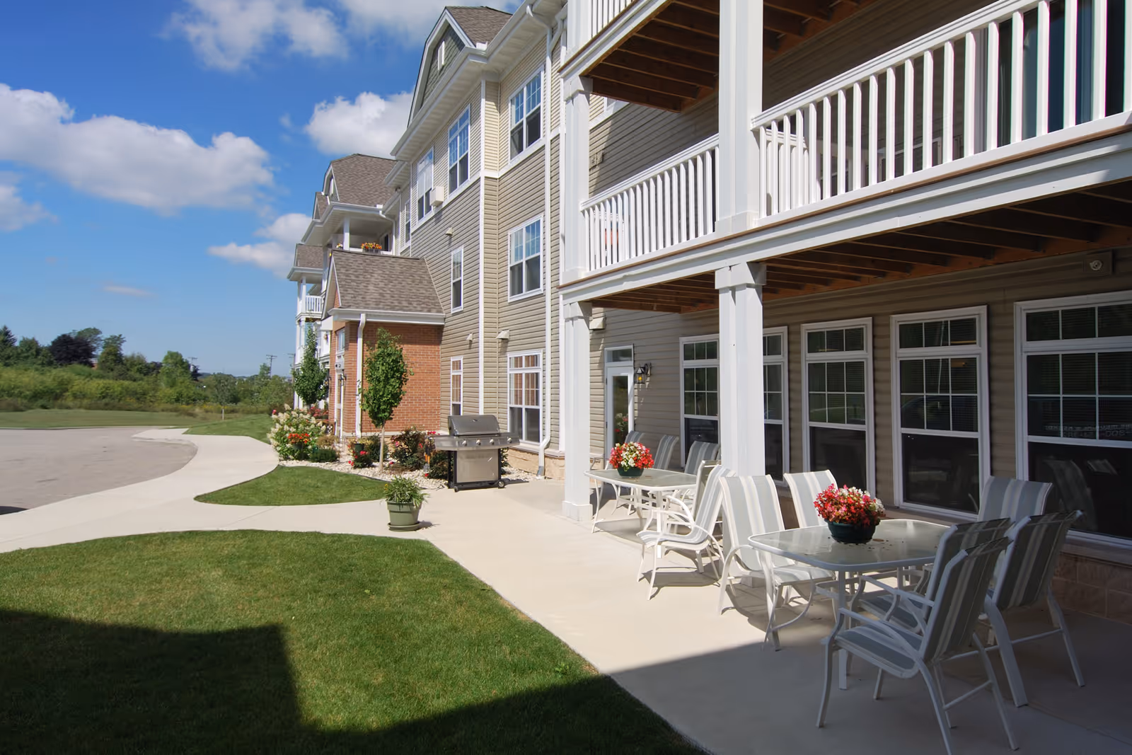 Outdoor patio with tables, chairs, potted flowers, and a grill in front of a multi-story senior apartment building with balconies and a lawn.