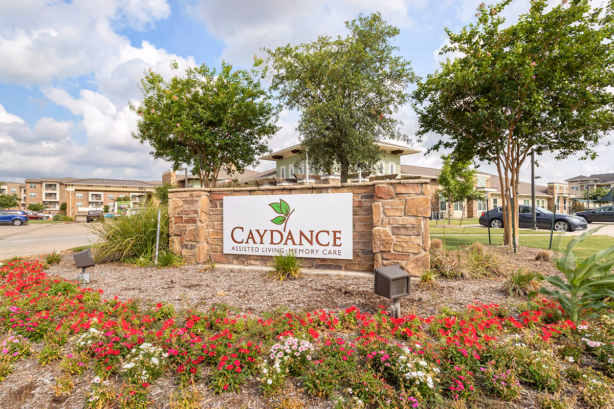 Outdoor view of the entrance sign for Caydance Assisted Living & Memory Care, surrounded by landscaped flowers, trees, and a stone wall with buildings and parked cars in the background under a partly cloudy sky.