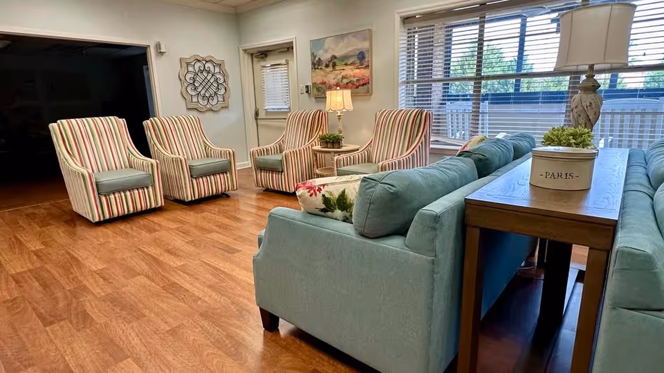 A cozy living room area with wooden flooring, featuring two light blue sofas with floral pillows, a wooden side table with a decorative container labeled 'PARIS' and a small plant, and four striped armchairs arranged near a wall with a door and a window with blinds. A lamp and wall art are also visible.