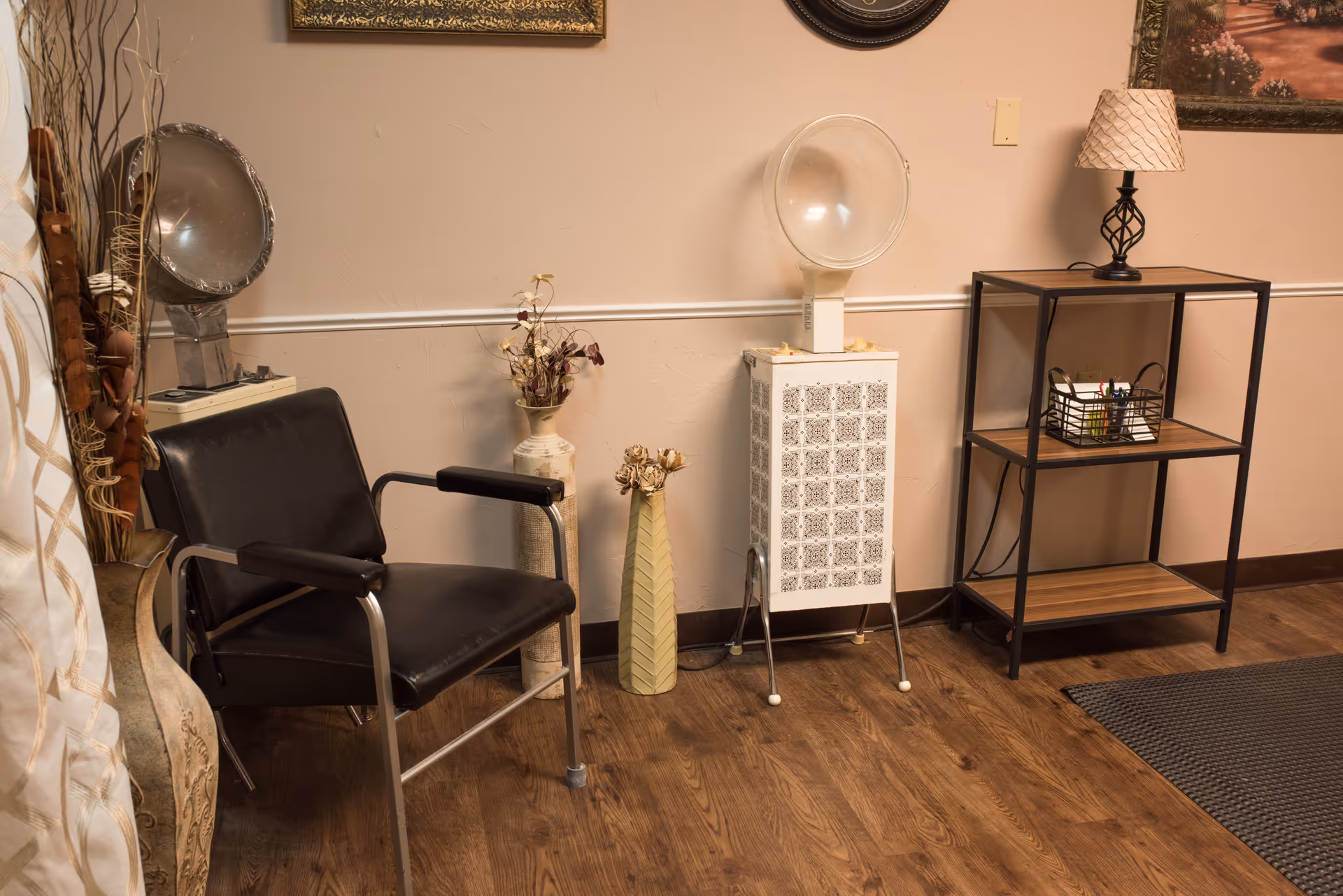 A cozy corner of a room with a black salon chair, two decorative vases with dried flowers, a vintage hair dryer, a small shelving unit with a lamp and a basket of pens, and framed artwork on the wall. The floor is wooden and there is a small rug partially visible.