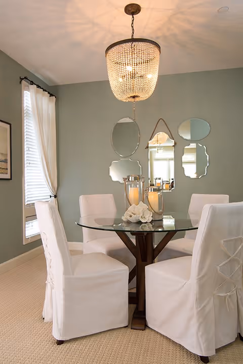 A dining room with a round glass table and four white slipcovered chairs. On the table are two large candle holders with lit candles and a decorative white coral piece. The walls are painted light green and decorated with five uniquely shaped mirrors. A large beaded chandelier hangs above the table, and a window with white curtains is on the left side.